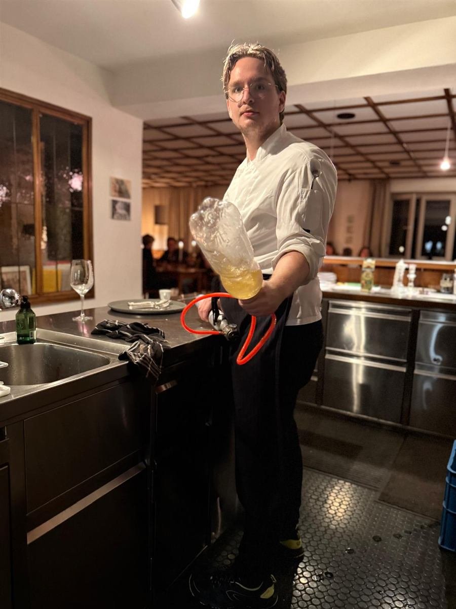 Koch mit Siphonflasche in der offenen Küche, im Hintergrund die Gäste